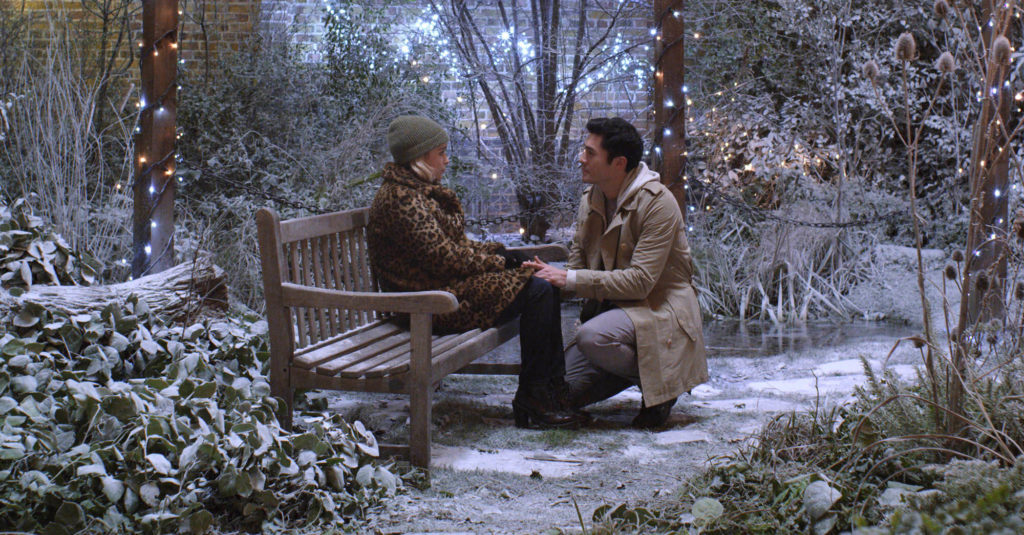 Emilia Clarke and Henry Golding talk on a bench in a snowy garden in LAST CHRISTMAS (2019)